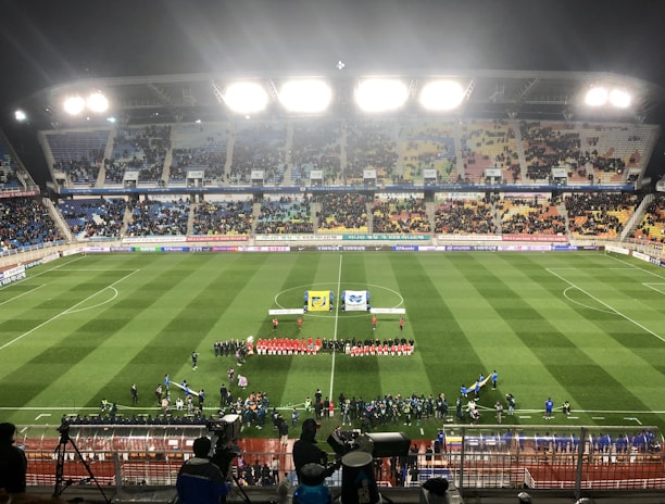 A large soccer stadium with bright overhead lights and a well-maintained green field. Teams are lined up in the center alongside officials, and there is a group gathered nearby, possibly for a pre-match ceremony. The stadium is filled with spectators wearing colorful clothing, and banners and advertisements are visible along the edges of the field. Cameras and officials are positioned on the sidelines to capture the event.