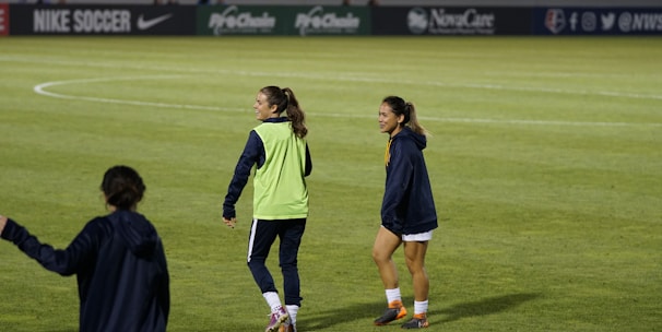 Two people are on a soccer field, wearing sports attire. One person is wearing a green vest, and the other is in a blue jacket and white shorts. The setting appears to be during a sports event, with adverts in the background and a partially filled stadium.