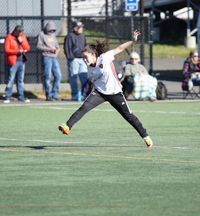 A person is mid-air on a sports field, appearing to be involved in an athletic activity. Several spectators are on the sidelines, some seated and others standing, with one individual capturing the event with a camera. The background includes a fence and various casual observers, some of whom are wearing winter clothing.