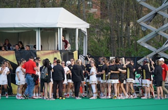 A group of athletes in sports uniforms is gathered on what appears to be a lacrosse field. They are standing in two groups, some wearing white uniforms and some wearing dark uniforms. There is a tent with people inside, possibly officials or commentators, overlooking the field in the background. Extensive greenery and trees are visible behind the tent.