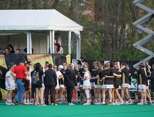 A group of athletes in sports uniforms is gathered on what appears to be a lacrosse field. They are standing in two groups, some wearing white uniforms and some wearing dark uniforms. There is a tent with people inside, possibly officials or commentators, overlooking the field in the background. Extensive greenery and trees are visible behind the tent.