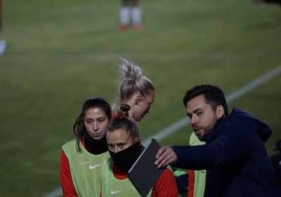 A group of individuals on a sports field are wearing green vests over red attire. One person is holding a tablet and seems to be discussing or showing something to the others. The atmosphere suggests a sports event, likely soccer, indicated by their clothing and the setting. The background is a grassy field.