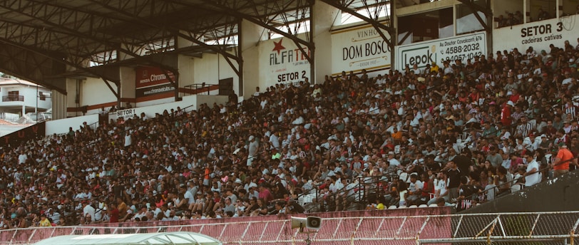 A crowded stadium with spectators filling the stands, observing an event. The stadium is covered and has multiple sections labeled with various advertisements visible on the walls. In front of the stands, a few people are gathered near an area with chairs and a white canopy.