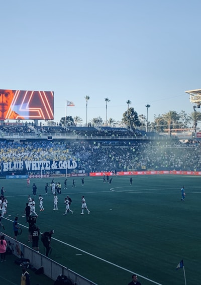 A large sports stadium filled with spectators. The field is occupied by soccer players, some walking and others scattered across the pitch. A massive screen displays a person, and flags fly on top of the stadium roof. The audience is dense, and the atmosphere seems lively with a visible banner showing 'BLUE WHITE & GOLD'.