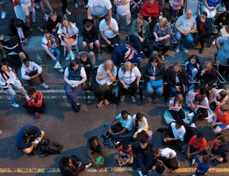 A large group of people gathered outdoors, watching a screen that is displaying a man. The crowd is diverse in age and appears interested and engaged, with many sitting closely together, some standing. Many are dressed casually, some in sports attire, and a few waving or holding flags. The event seems to have a communal, festive atmosphere.