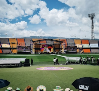 A sports stadium with a large stage set up on the field, hosting an outdoor event. Spectators wearing colorful hats sit in the foreground. The seating areas display vibrant colors, and a clear blue sky with scattered clouds is visible. Large screens display graphics related to the event.