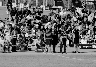 A large crowd of people is gathered, with some standing and others sitting on bleachers. Several individuals are wearing sports attire, and there is a field visible in the foreground with a marker indicating 50 meters. The scene appears to be a sporting event, with a diverse group of spectators.