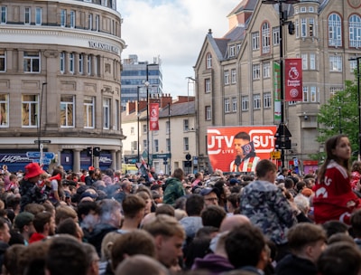 A large crowd of people, many wearing red and white, gather in a city square. They are watching an event broadcast on a large screen, which displays a man speaking into a microphone with logos suggesting a sporting event. Surrounding buildings are historical and commercial, with banners indicating a connection to the Premier League.
