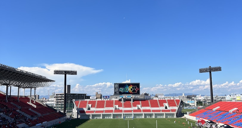 A large sports stadium with red and blue seating overlooks a green field with a goalpost at the center. A few people are scattered across the field, with some tents and a gathering of individuals on the right side. The sky is clear and blue with some clouds visible in the distance. A large screen or scoreboard is situated at the far end of the field.